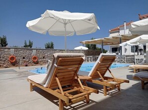 Two wooden lounge chairs with white cushions and a white umbrella, a stone wall, and a pool in the background. - Koukounari Apartments (Agistri)
