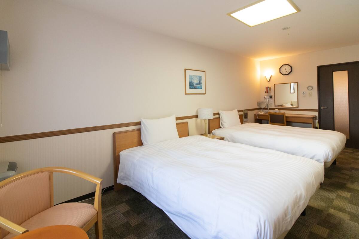Hotelkamer met twee bedden, een bureau, een stoel, een bedlampje, een klok en een lijst met een foto aan de muur.