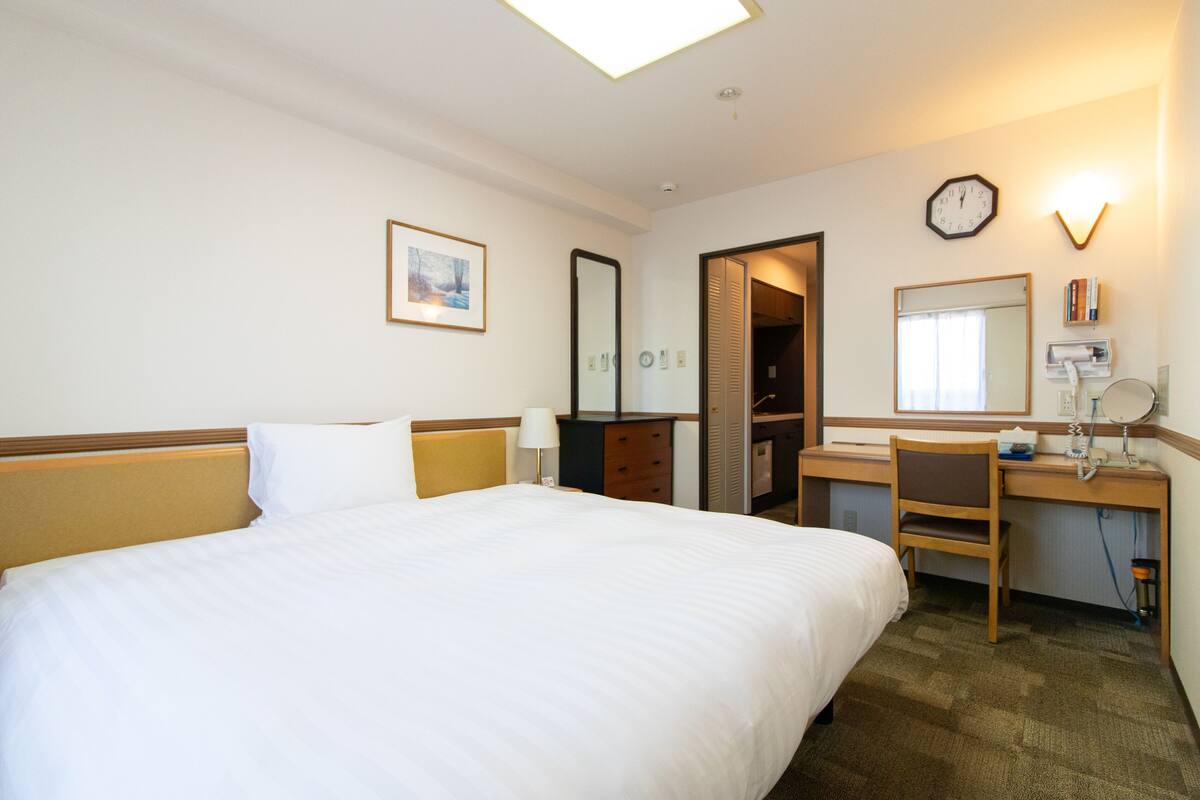Een hotelkamer met een groot bed, een bureau met stoel, een spiegel, een klok en een lijst met een foto aan de muur.