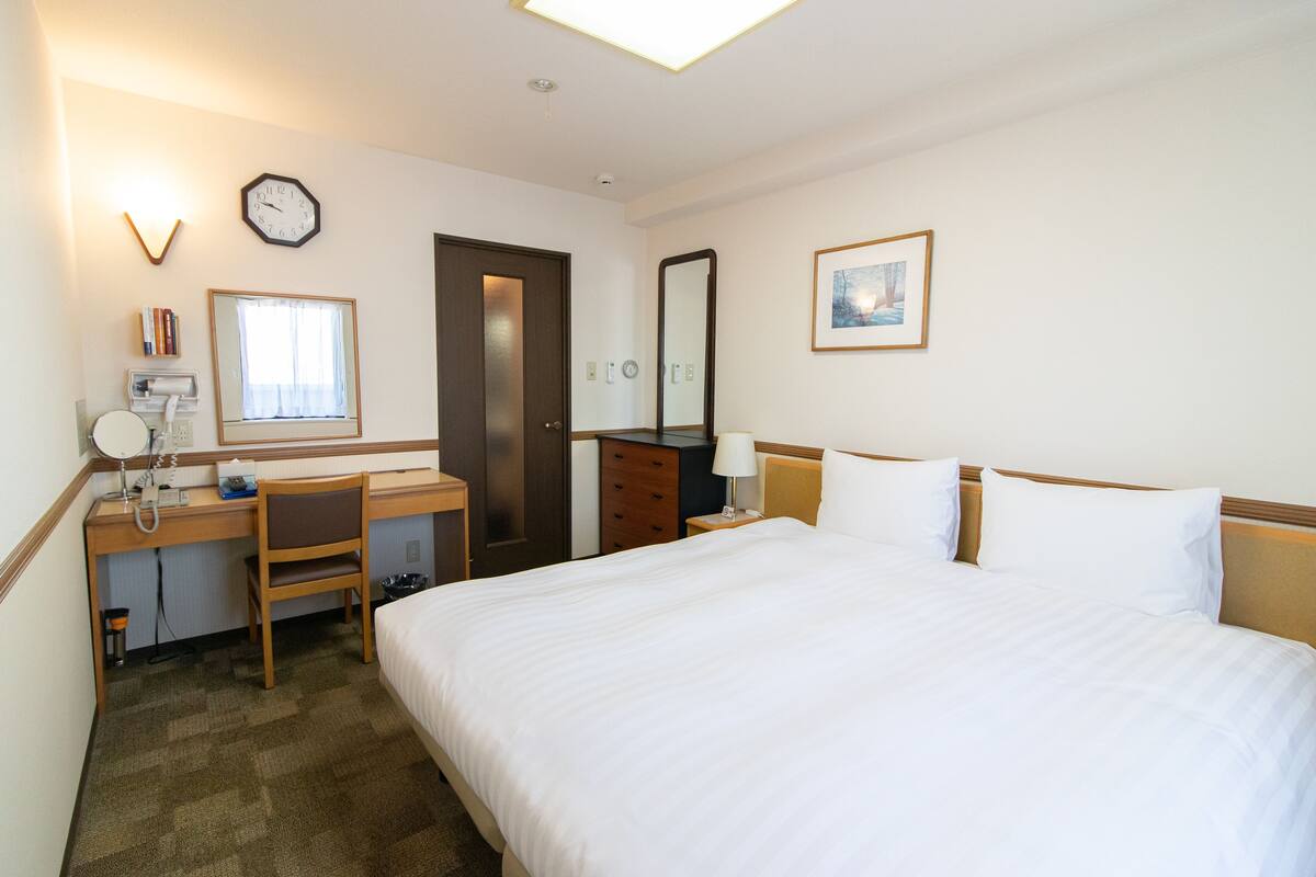 Hotelkamer met een bed, bureau, stoel, spiegel, klok, raam en een lijstje aan de muur.