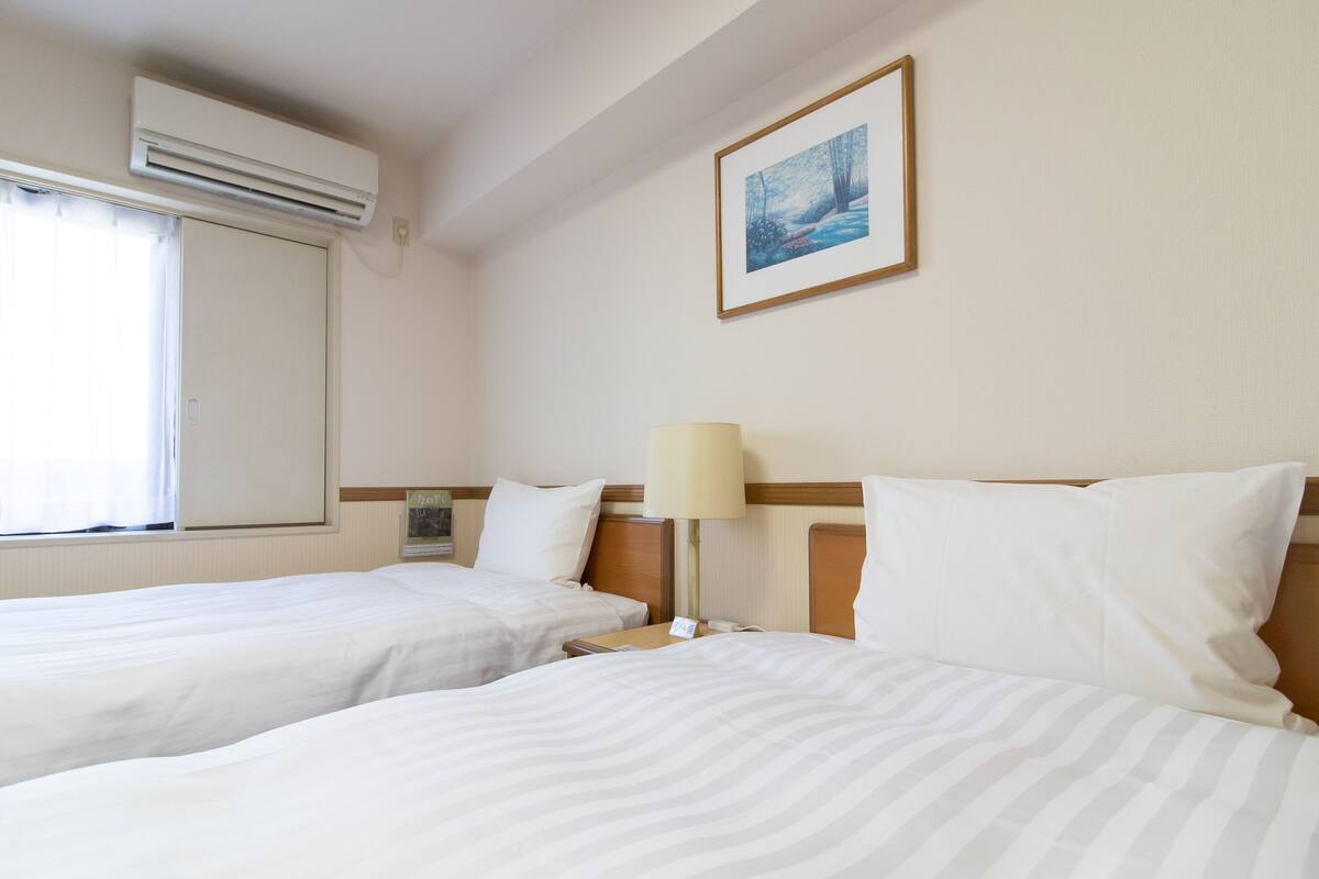 Een hotelkamer met twee bedden, een raam met gordijnen, een airconditioner, een bedlampje, een ingelijst schilderij aan de muur en een nachtkastje met een telefoon.