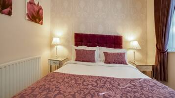 Une chambre d’hôtel avec un grand lit, deux tables de chevet, une tête de lit rouge, un papier peint à motifs et une fenêtre avec des rideaux.