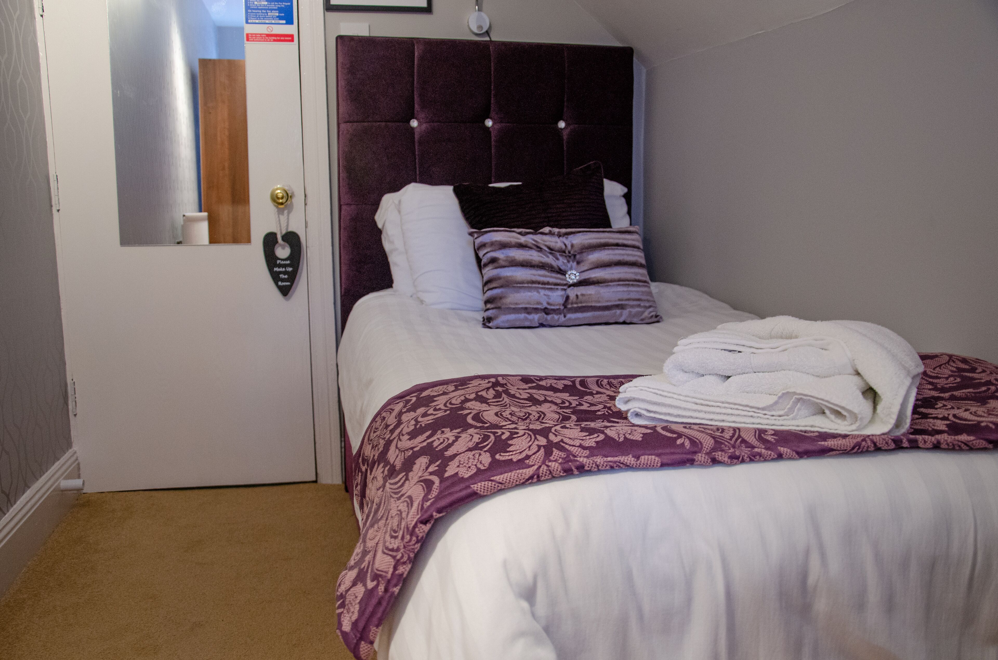 A neatly made bed with a purple and white bedspread, a folded white towel, and a heart-shaped decoration on the door.