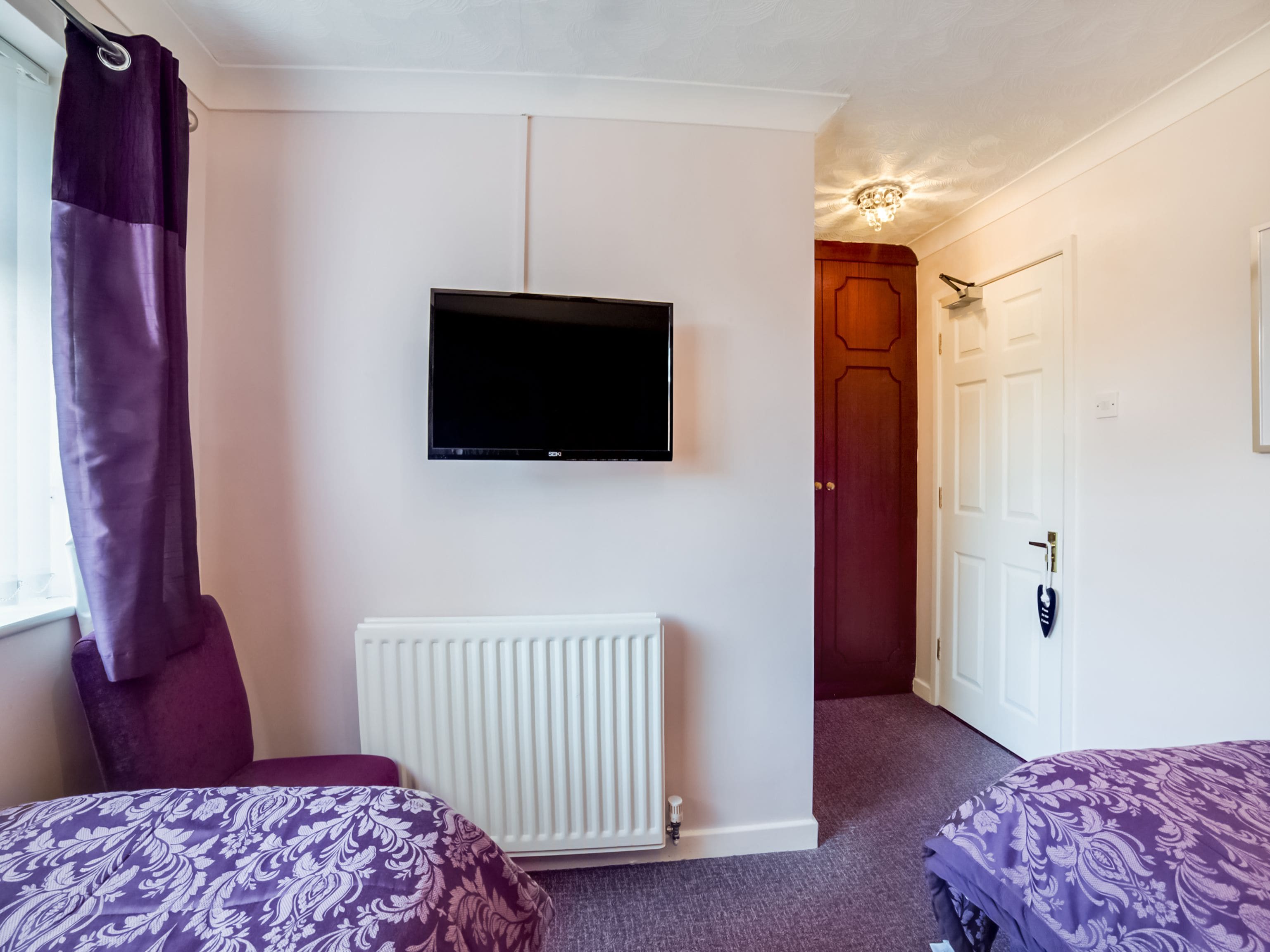 A bedroom with two beds, a TV, a radiator, and a door leading to another room.