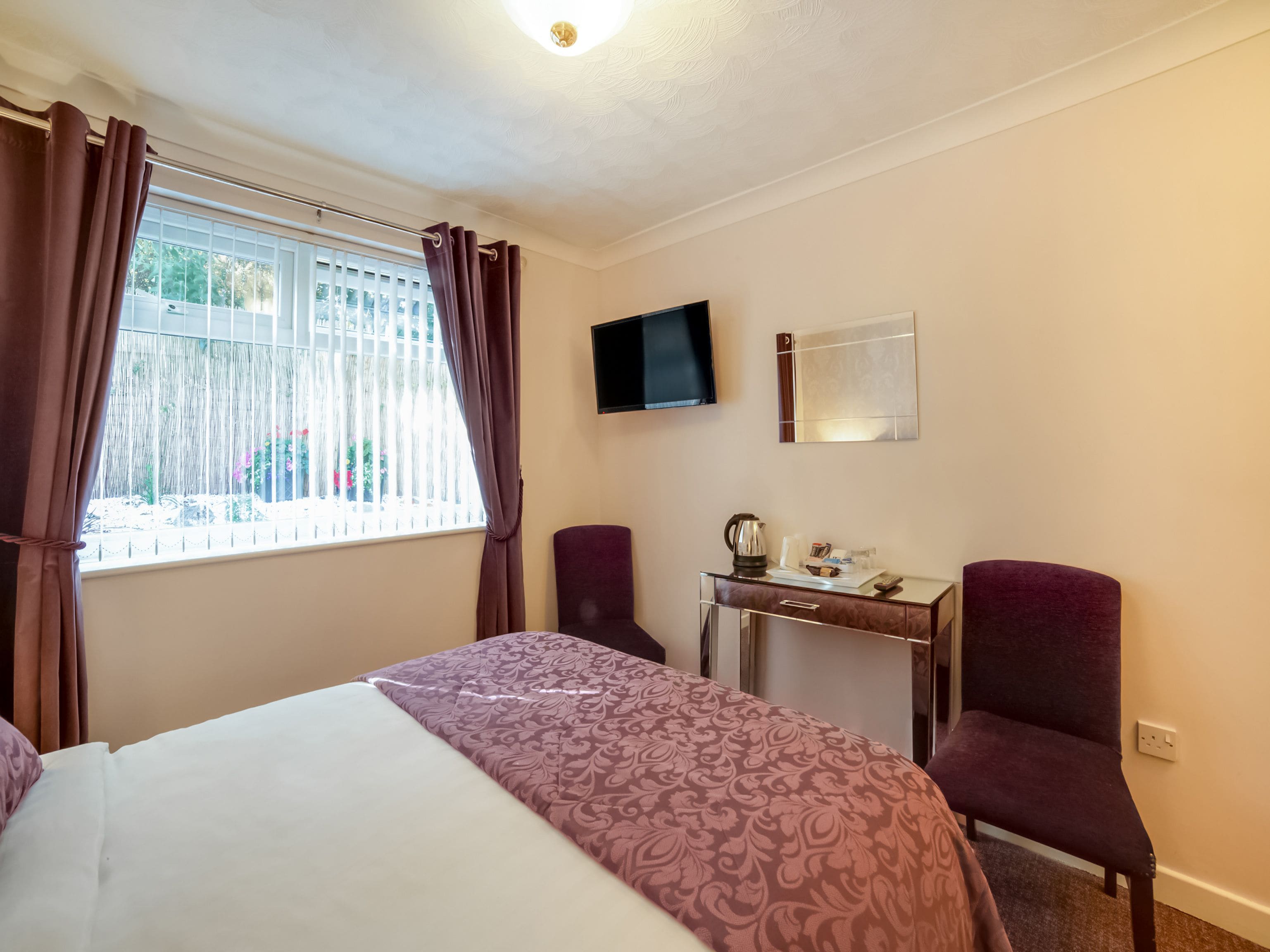 A hotel room with a bed, a desk with a kettle, two chairs, a TV, and a window with curtains.