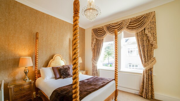 A four-poster bed with a wooden frame and a dark brown bedspread, a nightstand with a lamp, and a window with curtains. - Lemonfield Hotel (Sunderland)