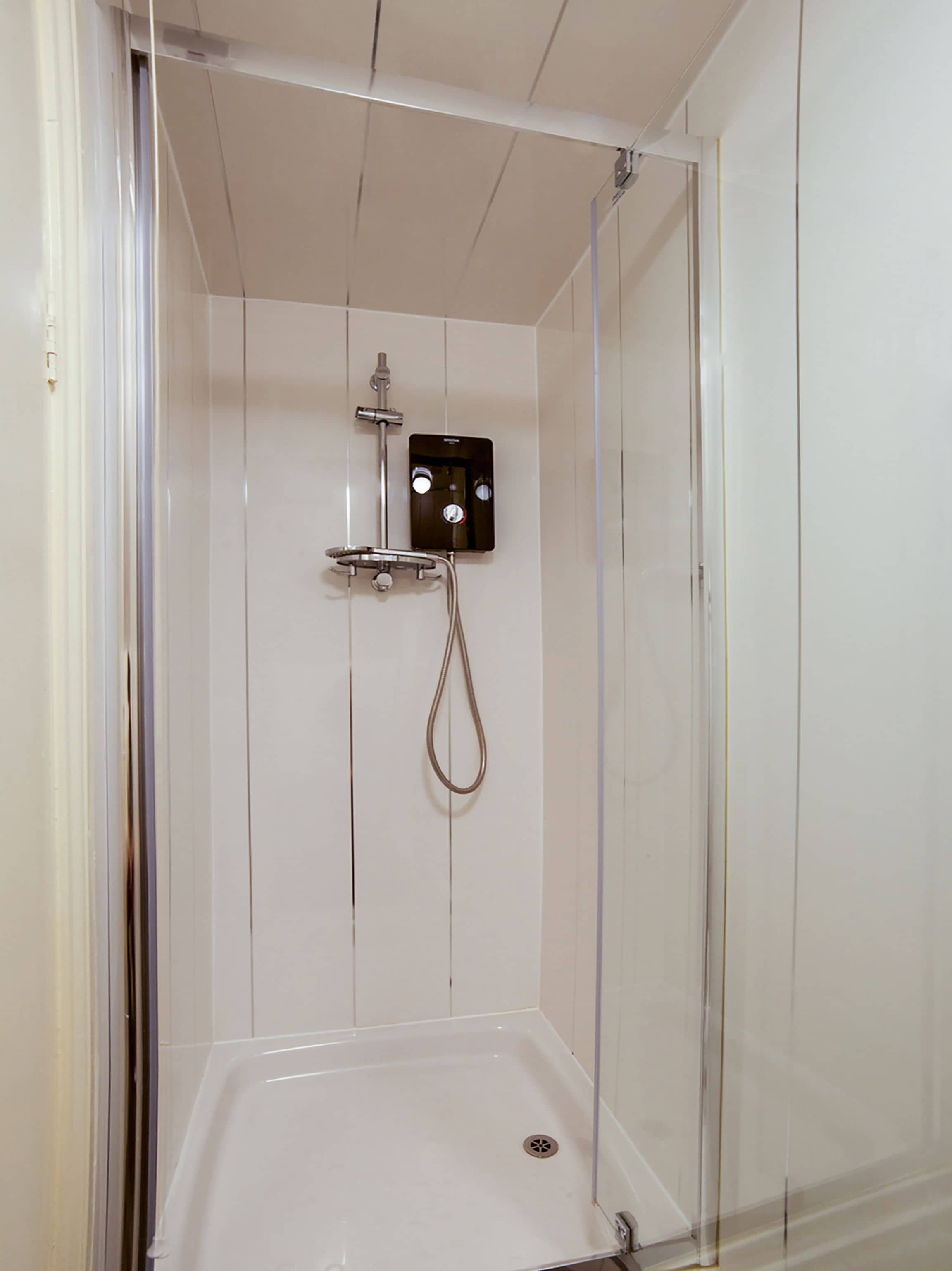 A small, enclosed shower area with a white tiled wall, a control panel, and a showerhead.