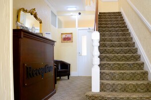 Zona de recepción con un mostrador de madera identificado como "Reception", un espejo, un cuadro enmarco y una escalera con alfombra estampada.