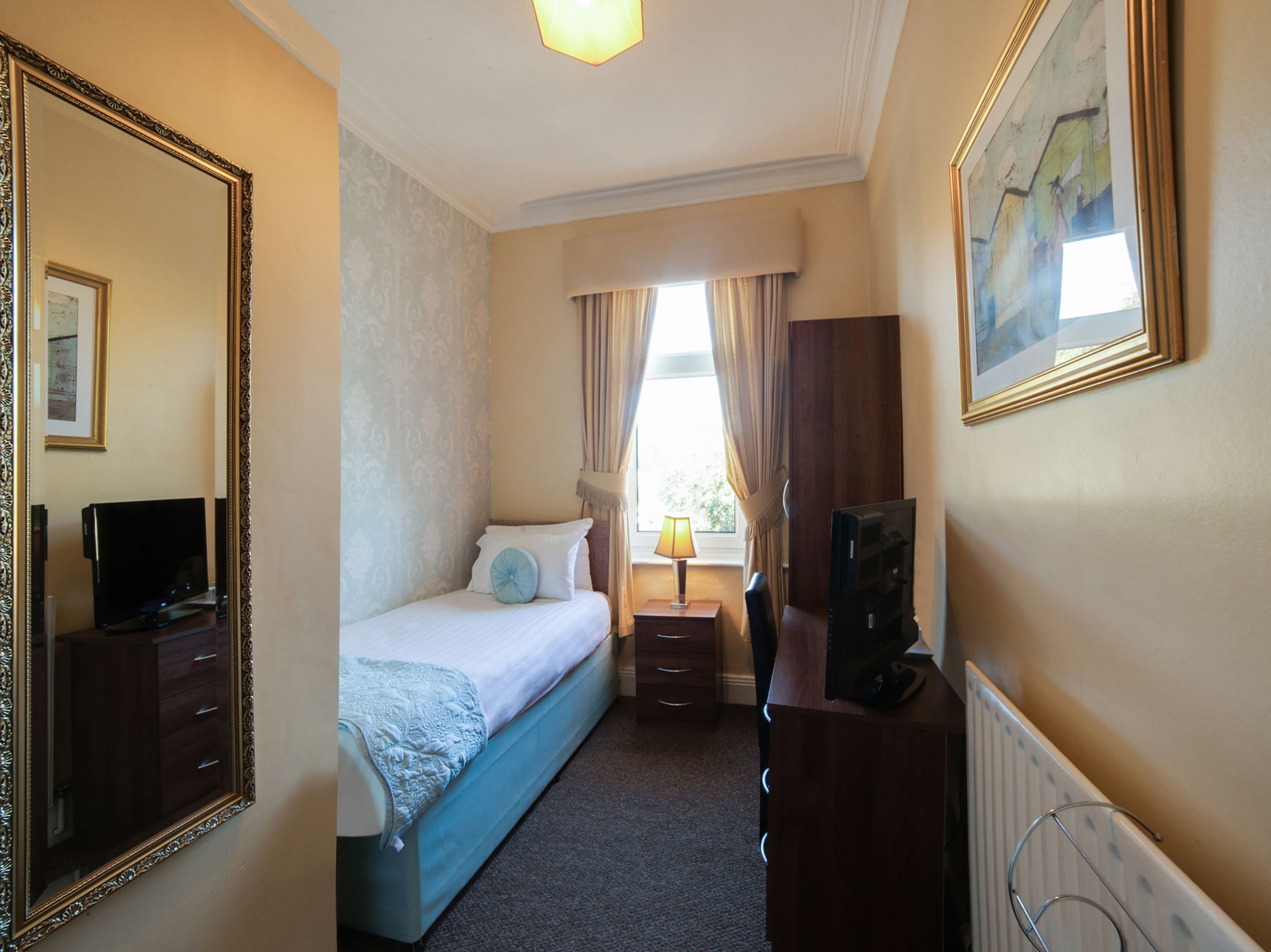 A hotel room with a bed, a desk with a computer, a television, a large mirror, and a window with curtains.