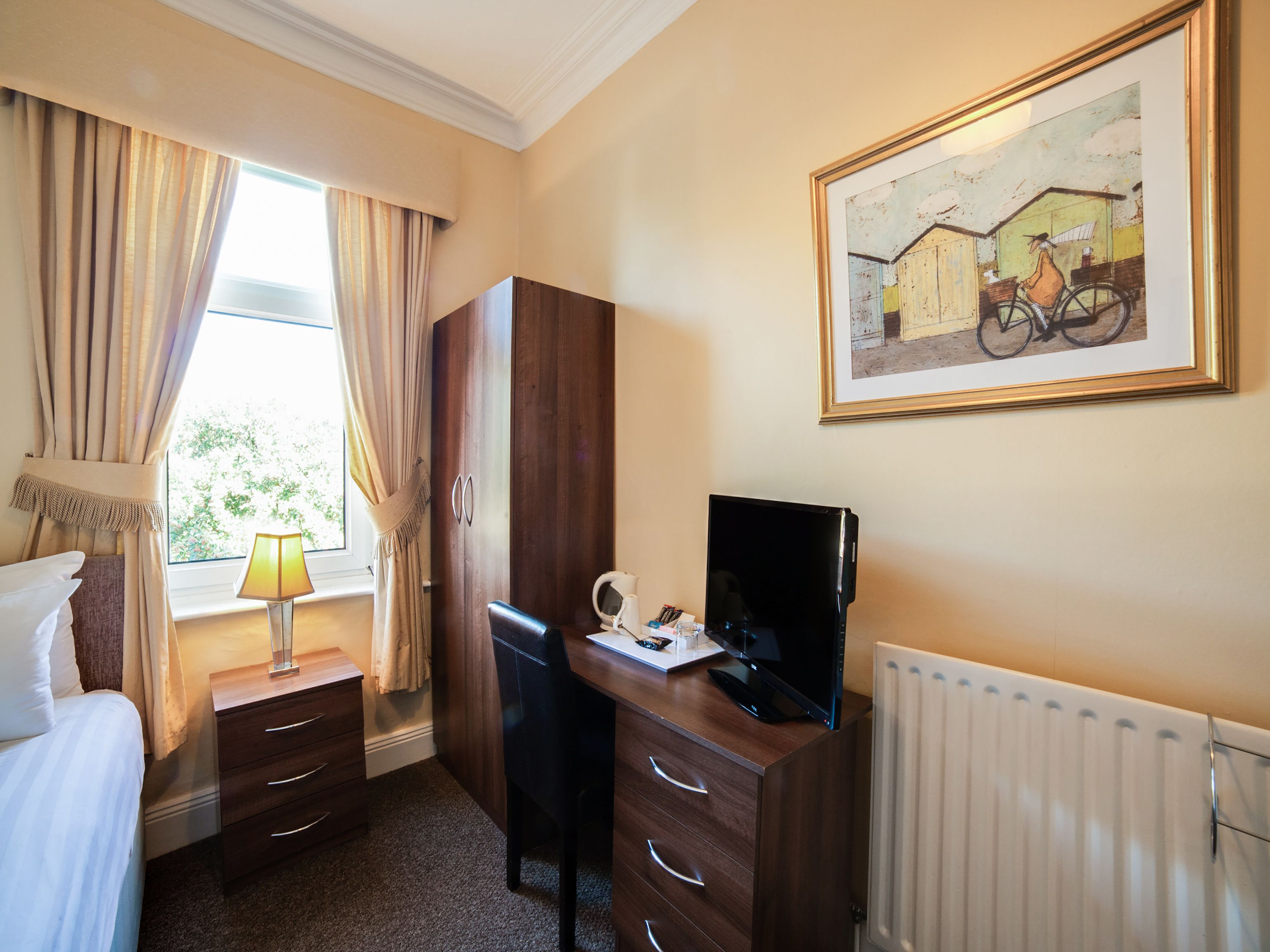 Una camera d'albergo con un letto, una scrivania con un computer, un termosifone e una finestra con tende.