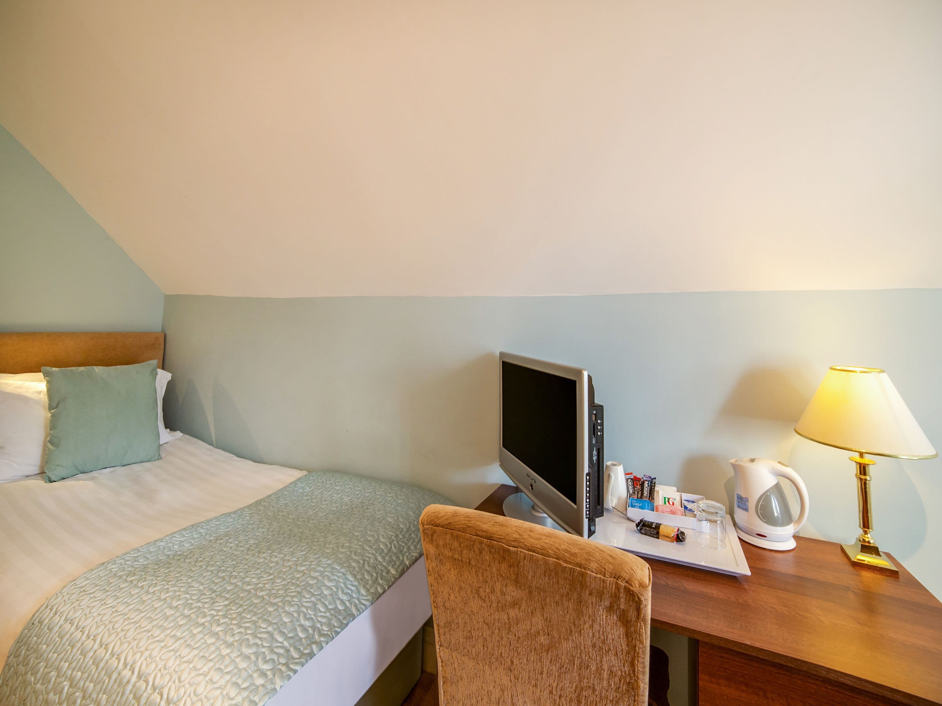 A hotel room with a bed, a desk with a computer, a lamp, and a kettle.