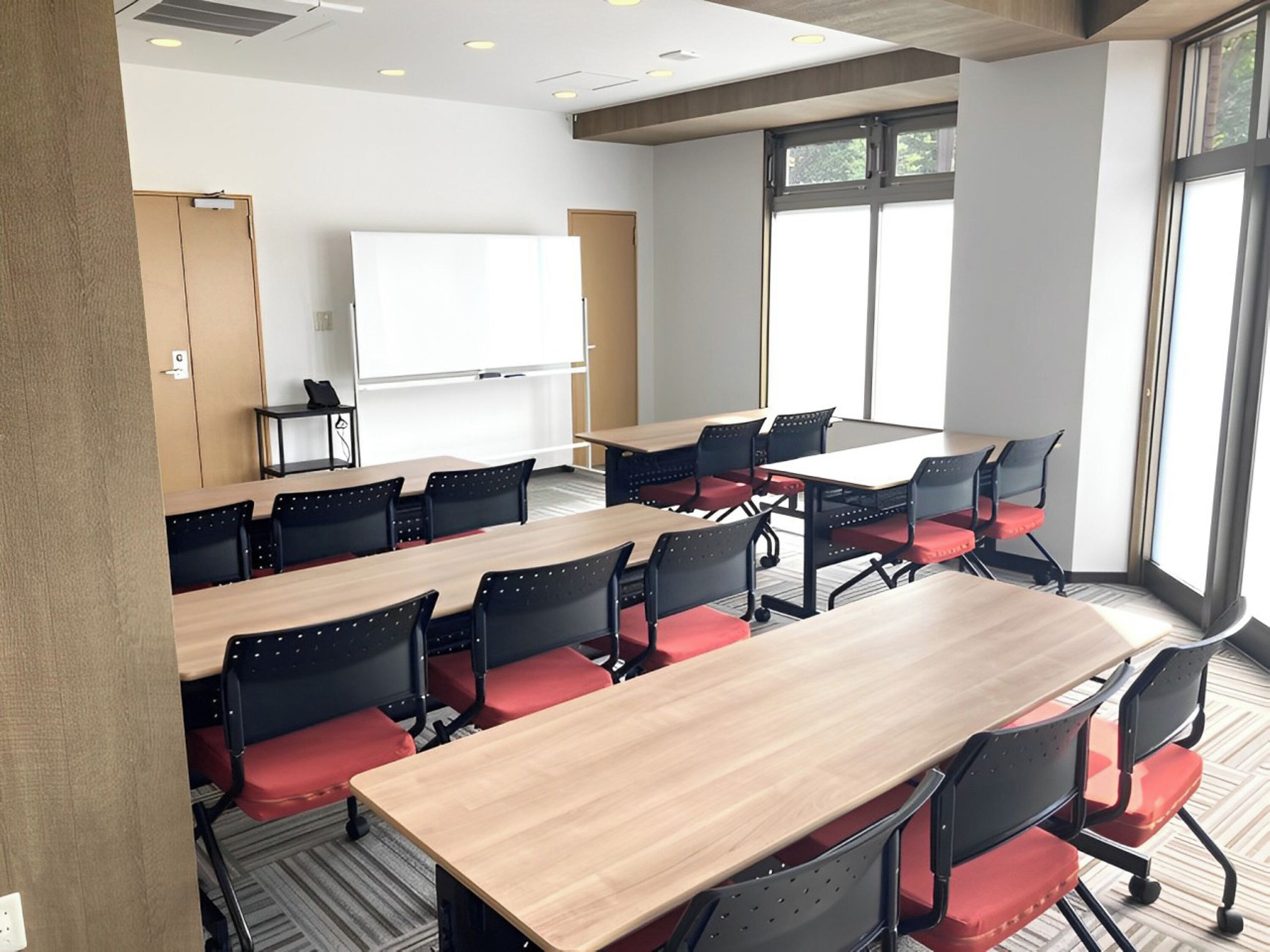 Uma sala de conferências com uma mesa grande, várias cadeiras, um quadro branco e janelas amplas.