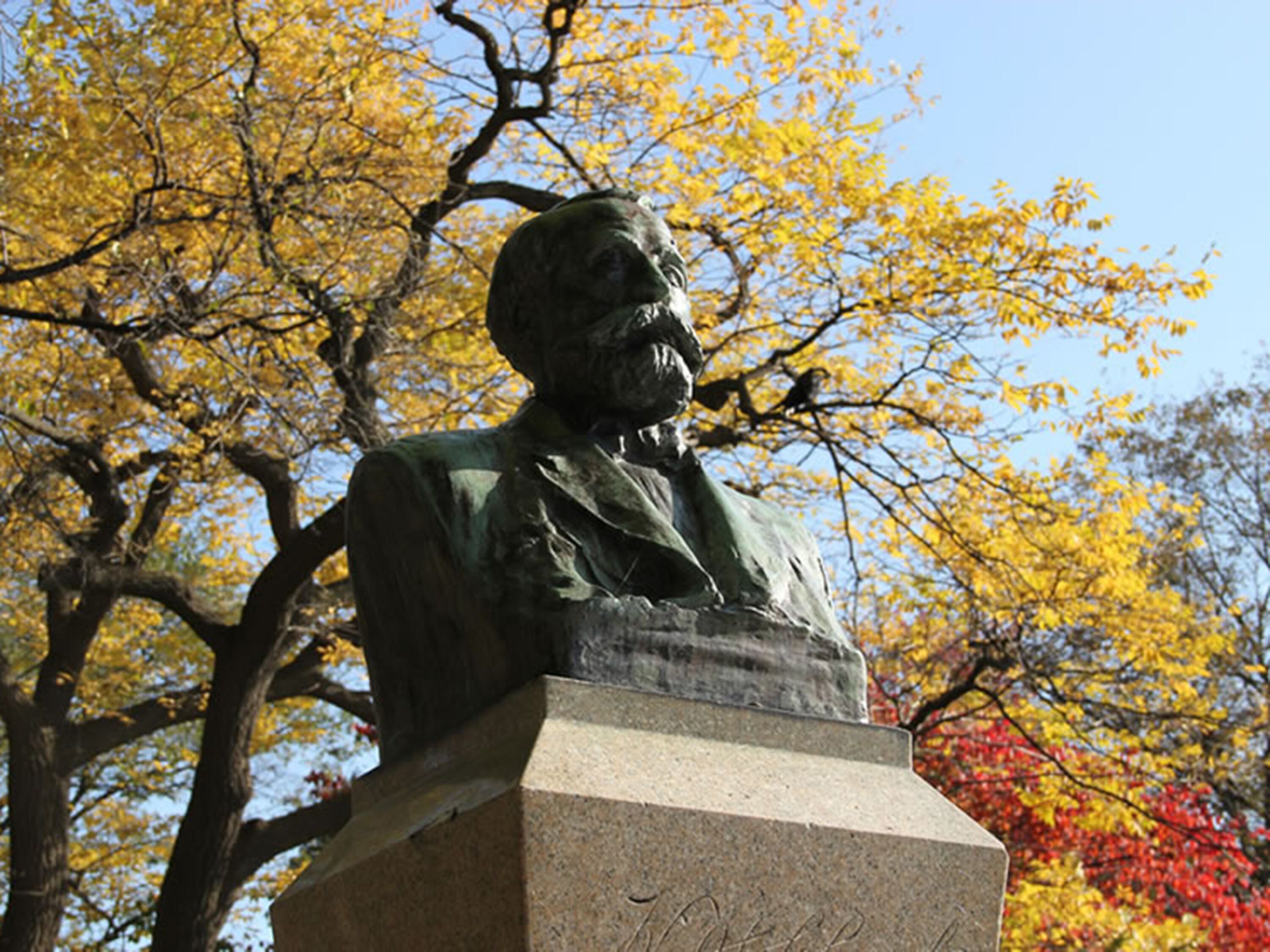 A statue of a man seated at a podium, surrounded by autumn-colored trees.