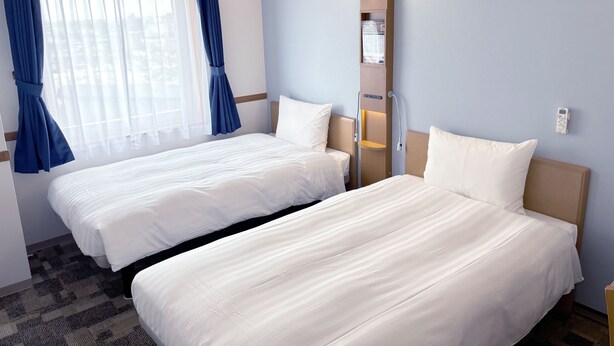 Habitación de hotel con dos camas, una ventana con cortinas azules, una lámpara fijada a la pared y un estante pequeño con una botella.