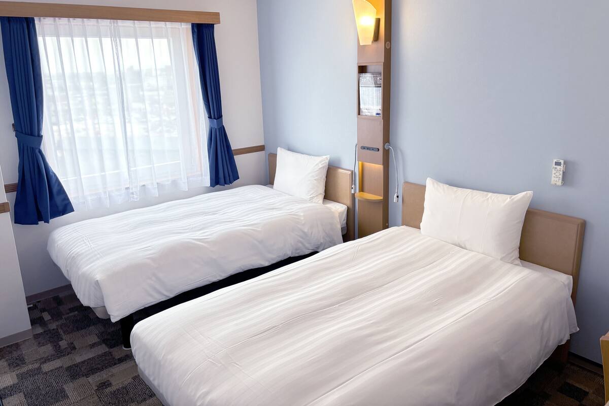 Habitación de hotel con dos camas, una ventana con cortinas azules, una lámpara fijada a la pared y un estante pequeño con una botella.