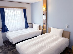 A hotel room with two beds, a window with blue curtains, a wall-mounted light, and a small shelf with a bottle. - Toyoko Inn Hokkaido Tokachi Obihiro Ekimae (Obihiro)