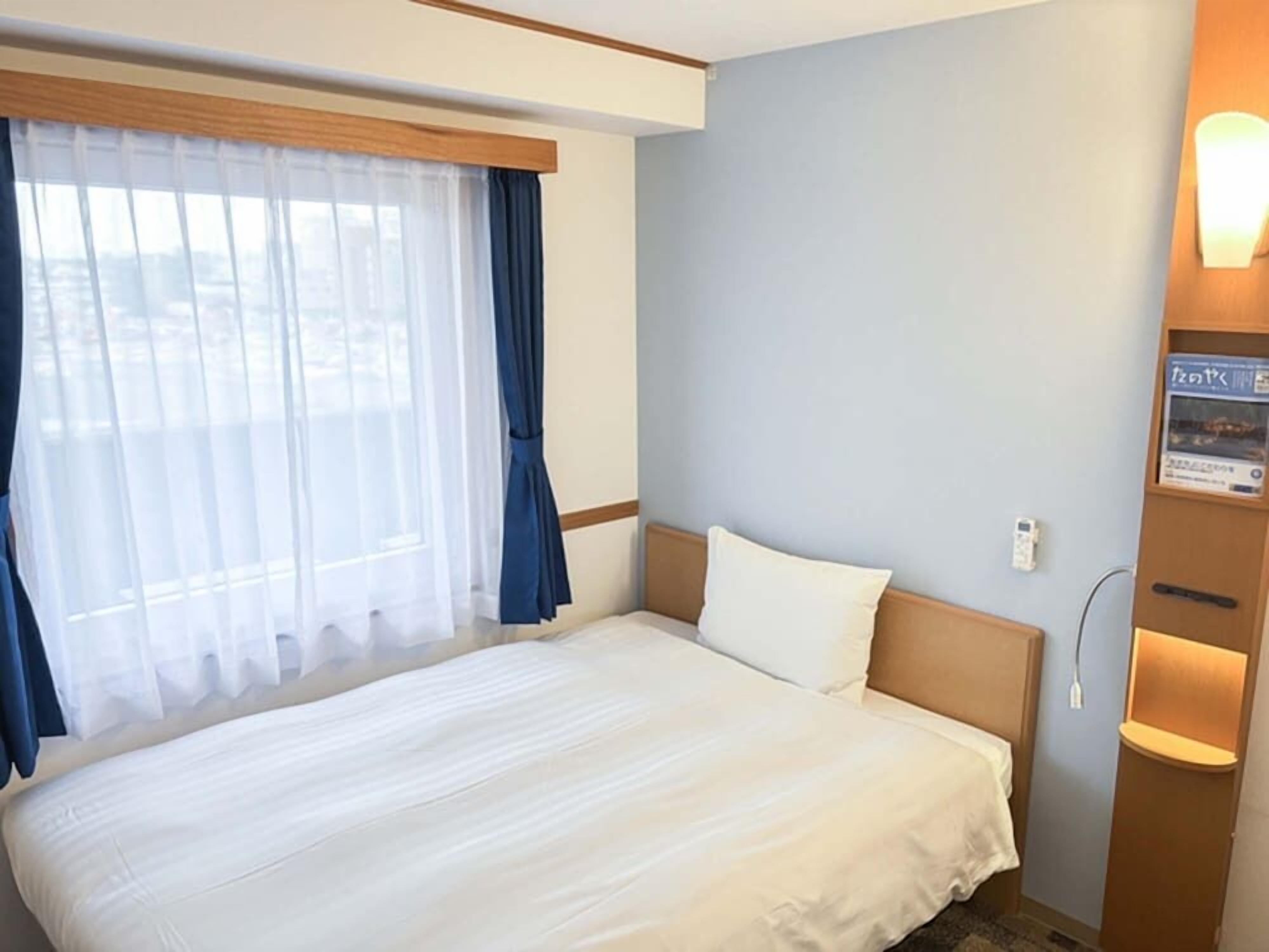 Habitación de hotel con una cama grande, una ventana con cortinas, una lámpara de pared y un mueble pequeño con luz.