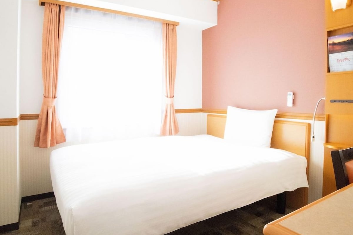 Una habitación de hotel con una cama individual, una mesita de noche de madera, una pequeña mesa y una ventana con cortinas.