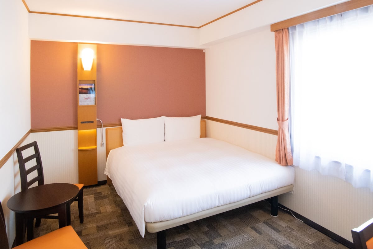 Et hotelværelse med en enkelt seng, et lille bord, en stol, et natbord og en lampe monteret på væggen.