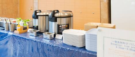 Disposição de buffet de café da manhã com diversos recipientes de comida, máquina de café e pilhas de pratos.