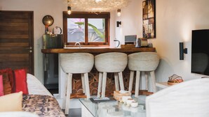 A compact kitchen with a wooden counter, three stools, a hanging light fixture, and a window with a view of greenery. - Villa LeoNora (Ubud)