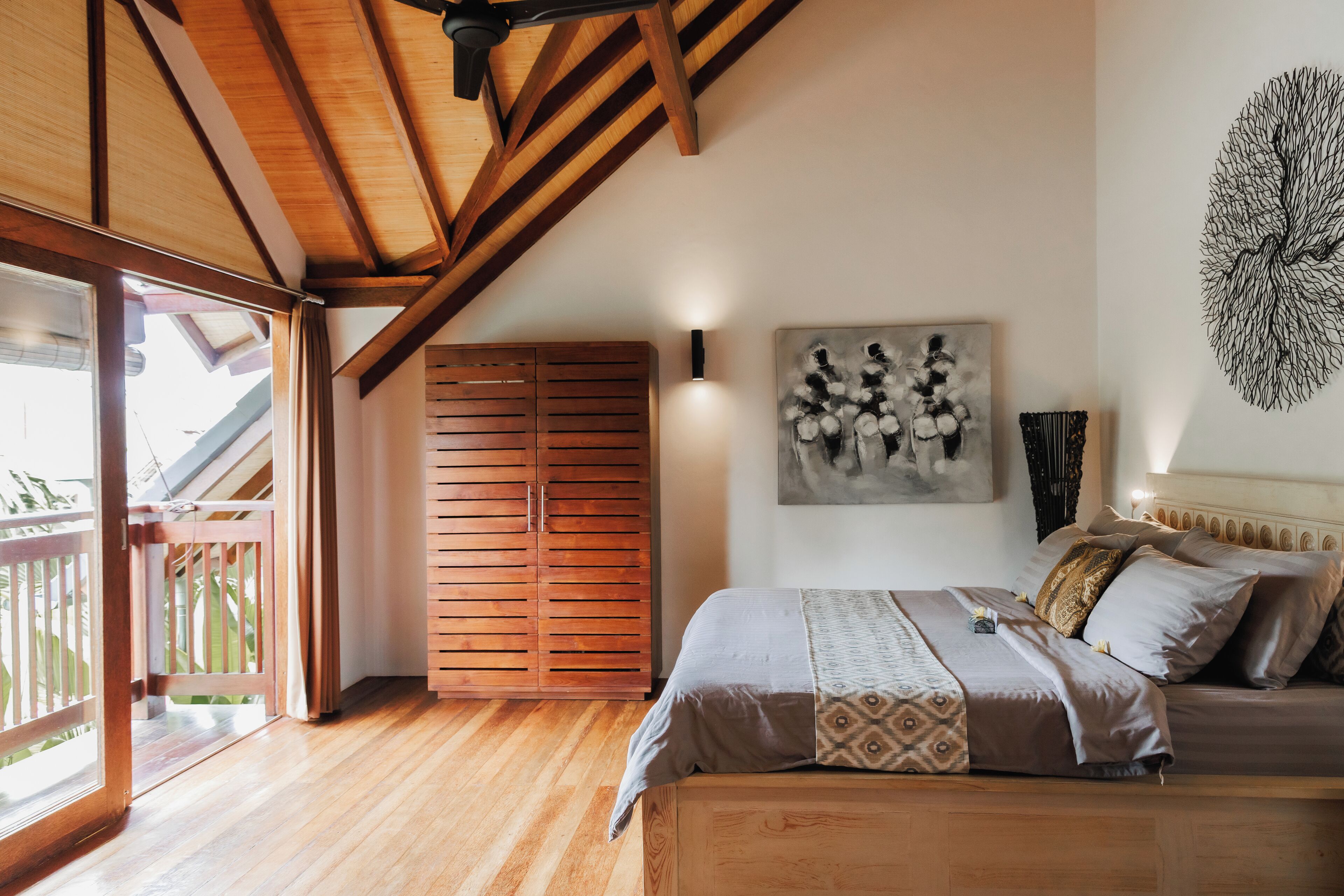 A bedroom with a wooden ceiling, a bed with patterned bedding, a wall-mounted artwork, and a wooden wardrobe.