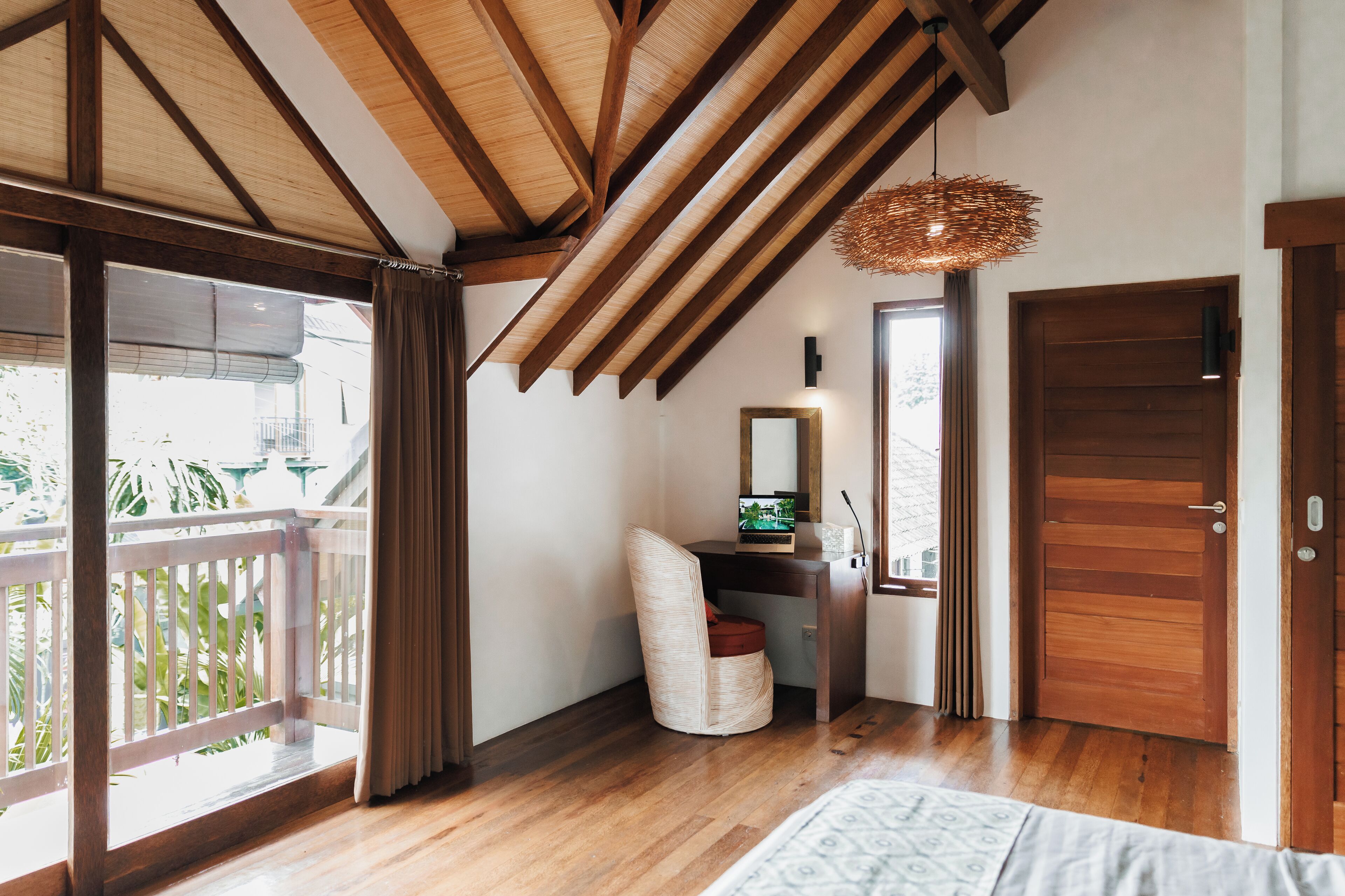 A room with a wooden ceiling, a desk with a computer, a chair, a bed, and a door.
