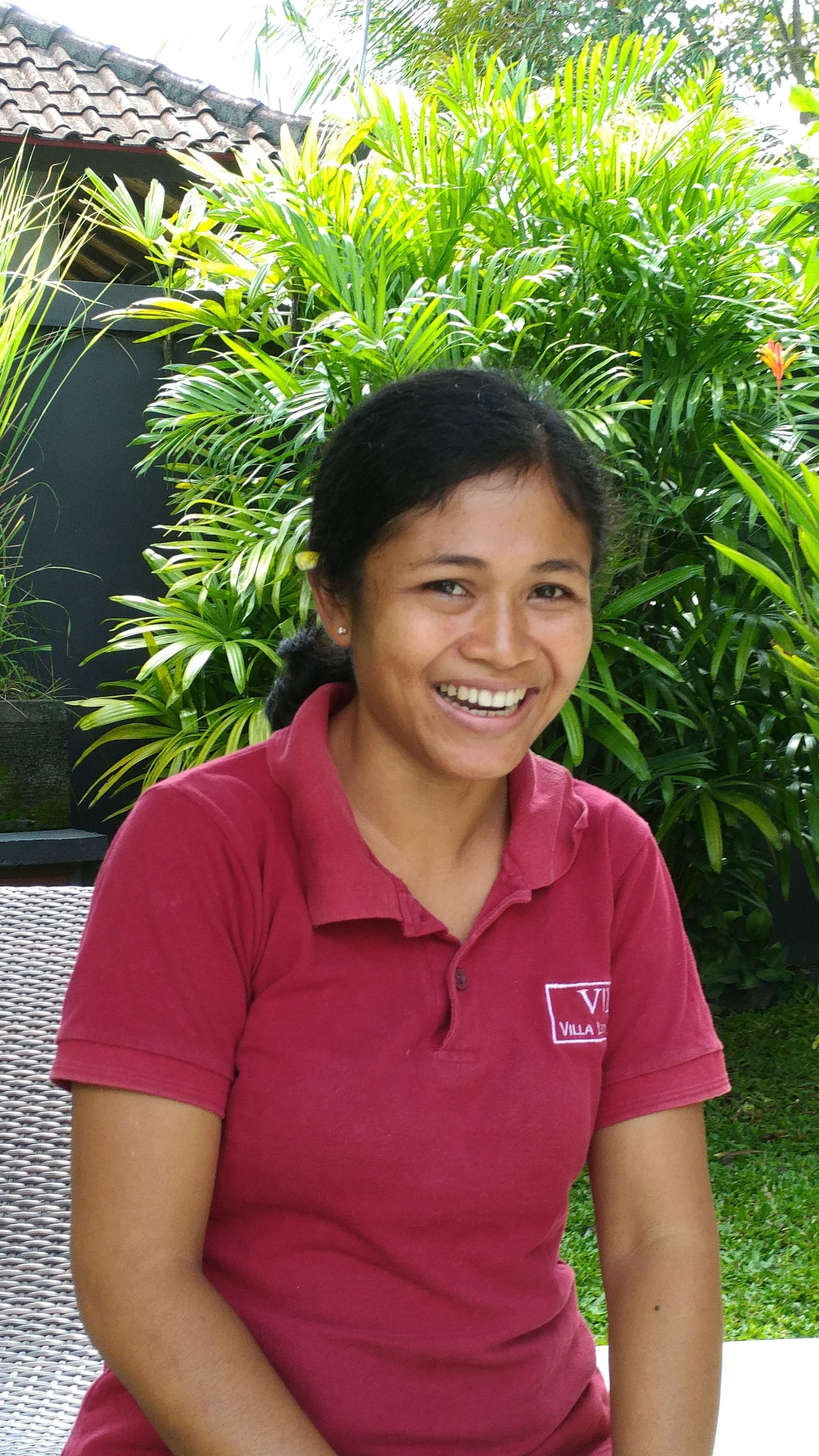A person wearing a red polo shirt with a logo, sitting outdoors with lush green plants and a tiled roof in the background.