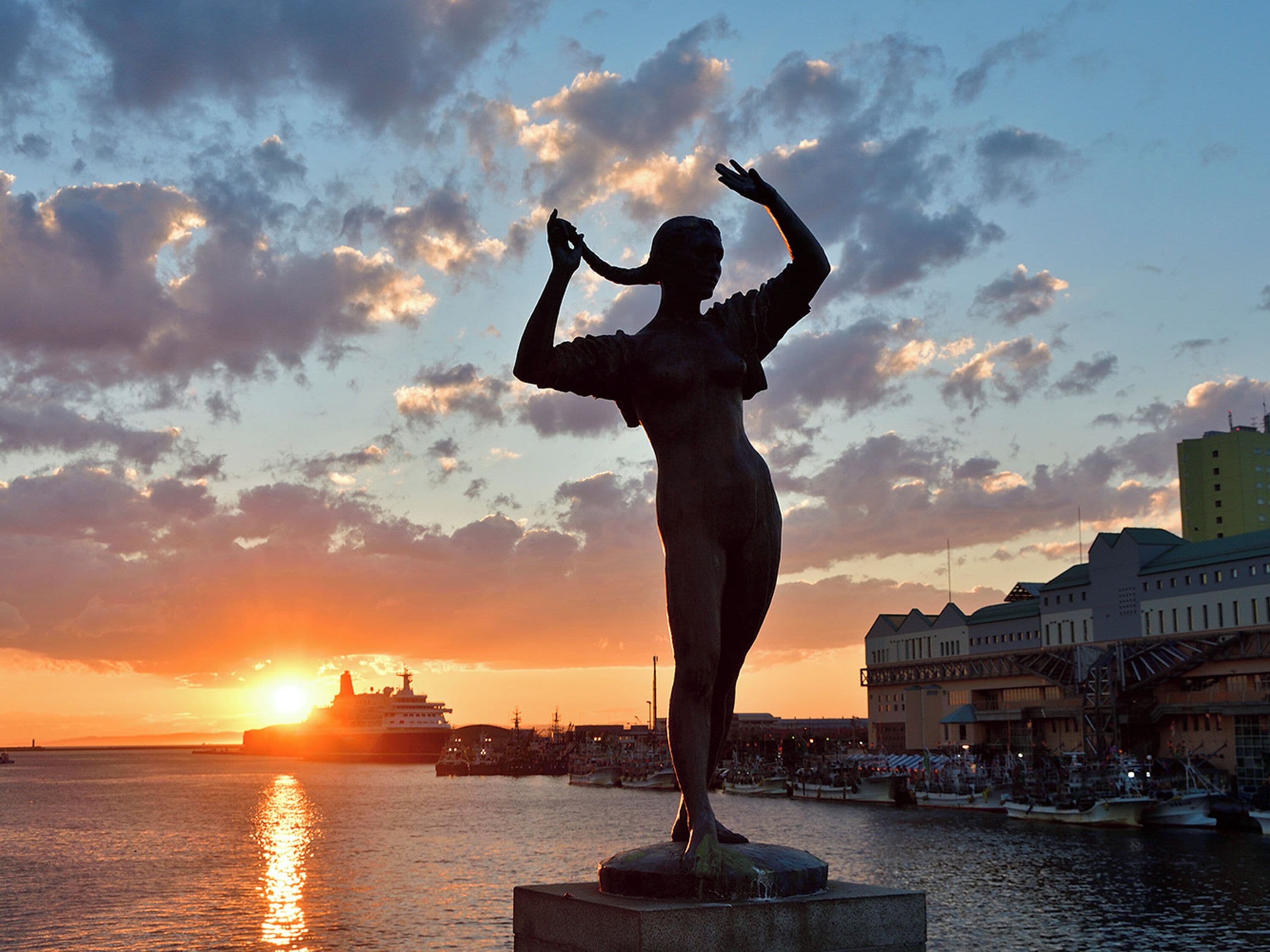 Estátua de uma mulher com os braços levantados, próxima a uma orla com o pôr do sol ao fundo.