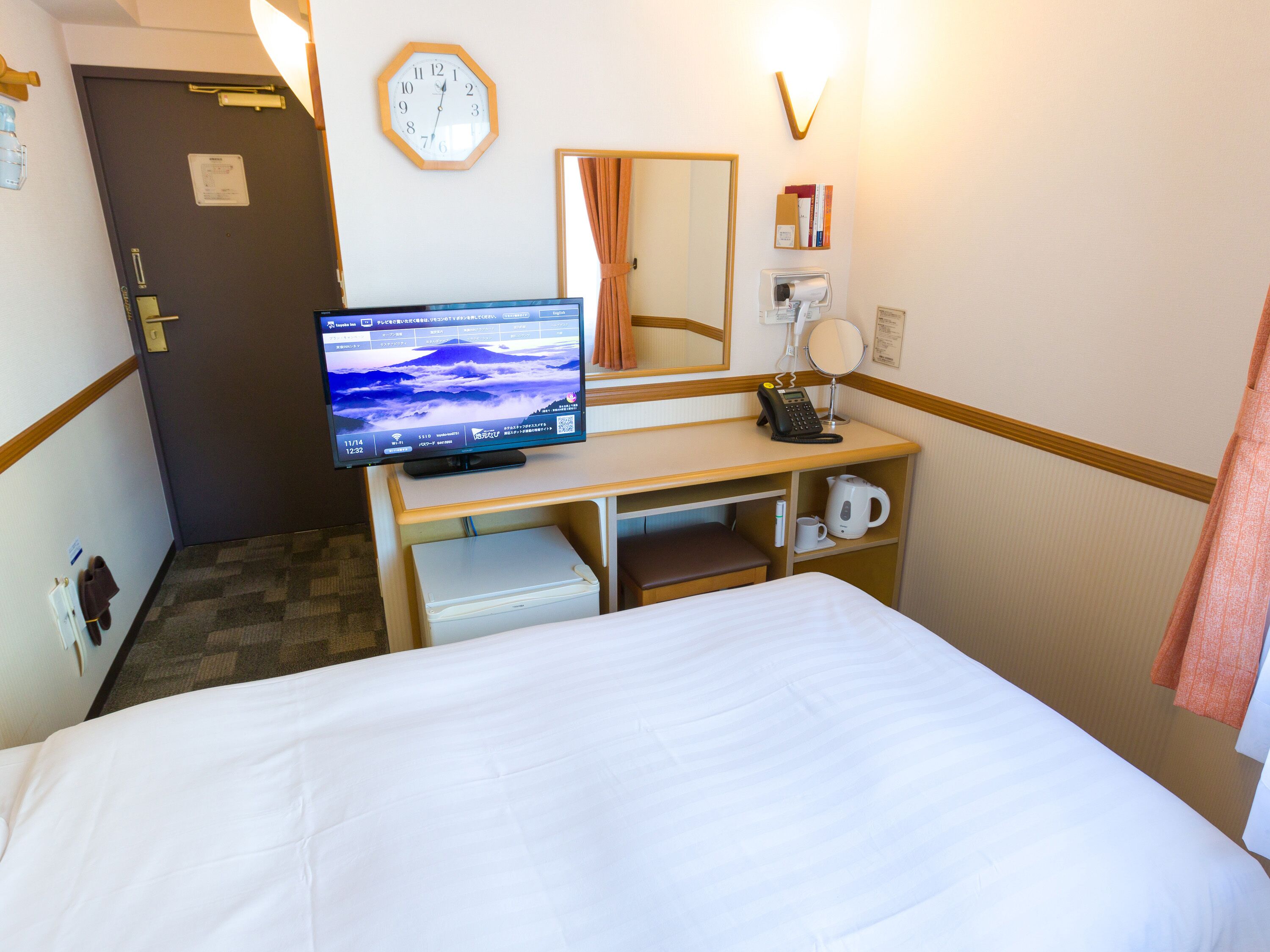 A hotel room with a bed, a desk with a computer monitor, a telephone, a kettle, and a clock on the wall.