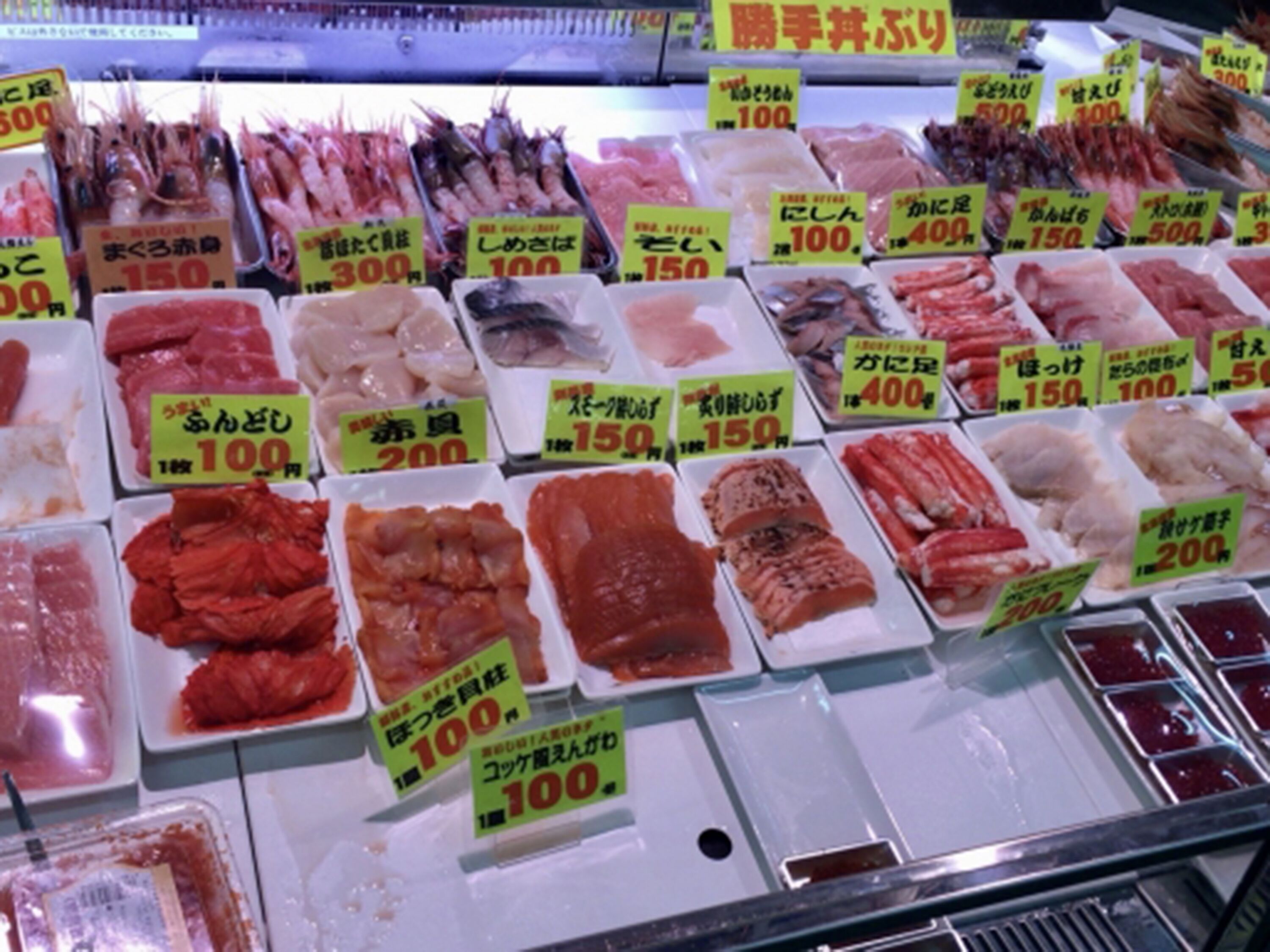 Une variété de produits de la mer est exposée dans une section réfrigérée d’un marché, et les étiquettes indiquant les prix sont bien visibles.