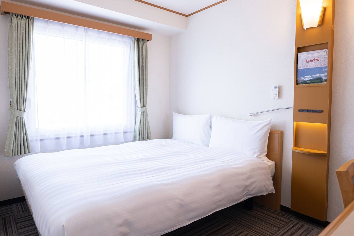 A hotel room with a large bed, a bedside table, a wall-mounted shelf with a book, and a window with curtains.