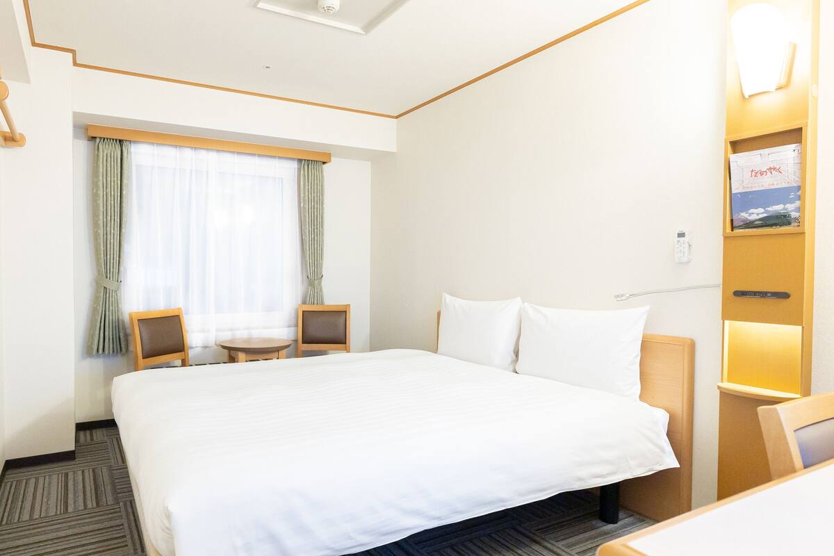 A hotel room with a large bed, a small table, two chairs, a window with curtains, and a wall-mounted TV.