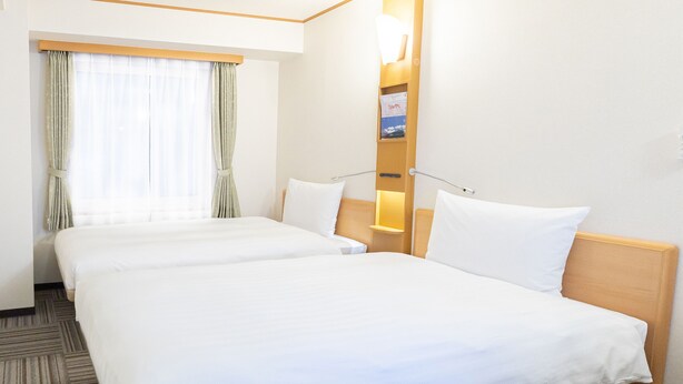 Ein Hotelzimmer mit zwei Betten, einem Nachttisch, einer Lampe, einem Fenster mit Vorhängen und einem an der Wand befestigten Regal mit einem gerahmten Bild.