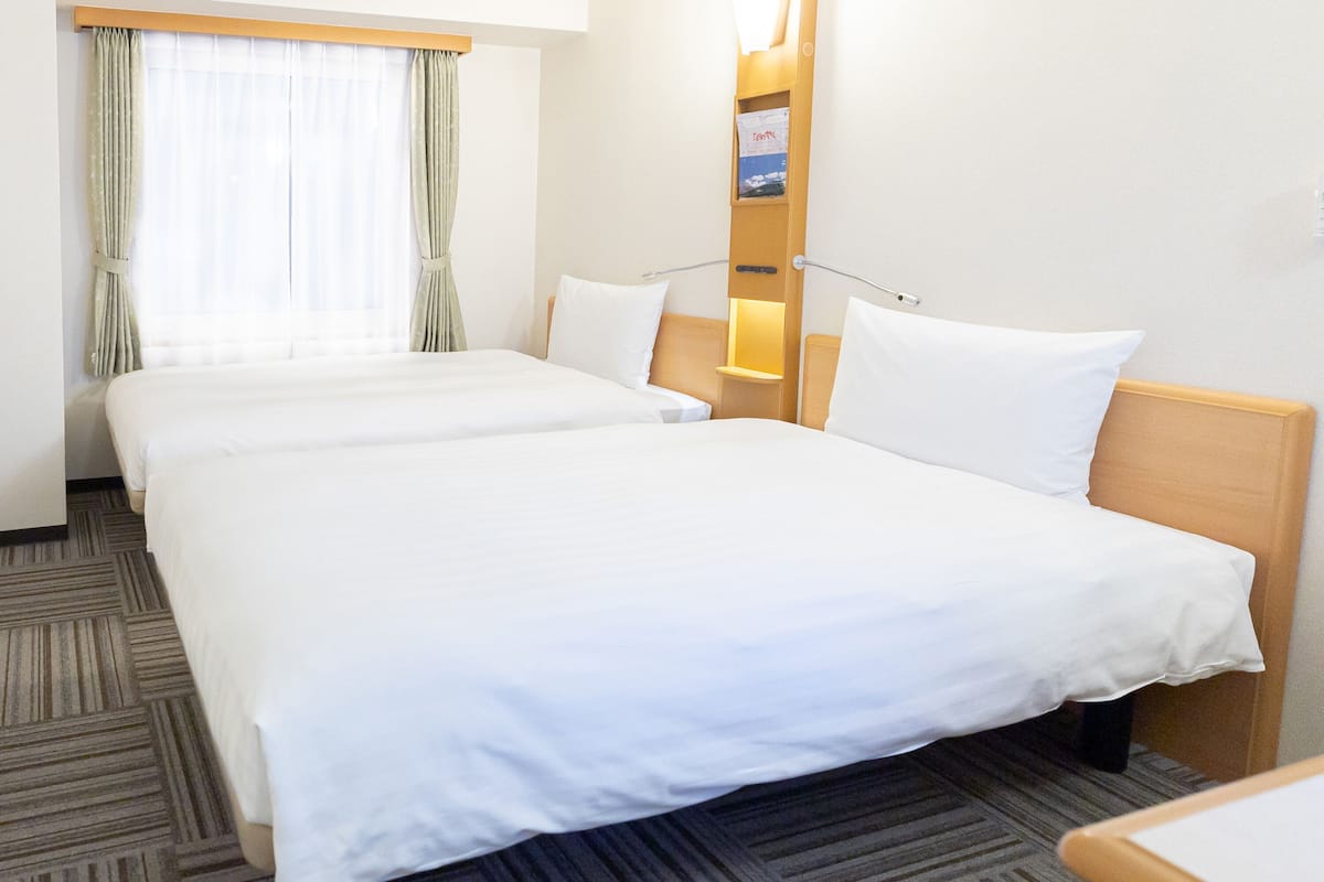 A hotel room with two beds, a nightstand, a lamp, a window with curtains, and a wall-mounted shelf with a framed picture.