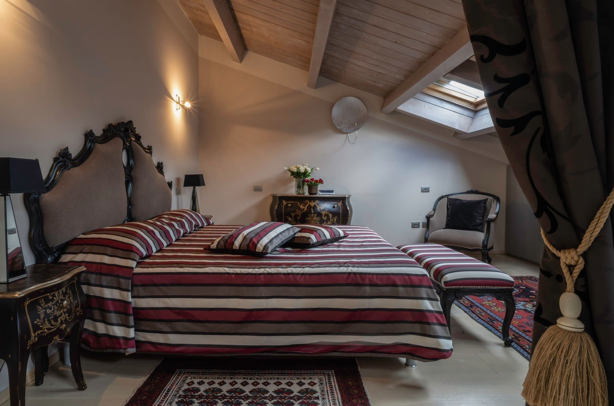 Ein Schlafzimmer mit einem großen Bett, einem Nachttisch, einem Sessel, einem kleinen Tisch mit Blumenvase und einem Deckenventilator.