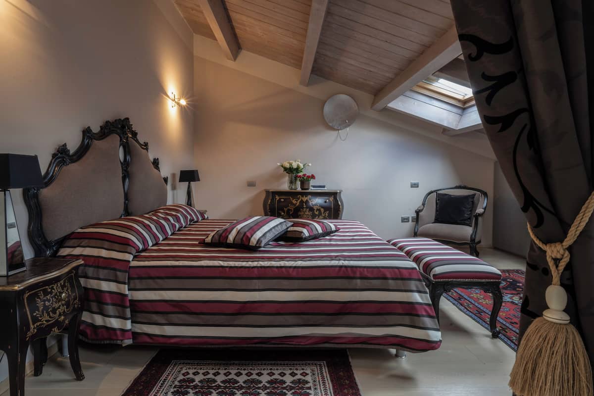 A bedroom with a large bed, a side table, a chair, a small table with a vase of flowers, and a ceiling fan.