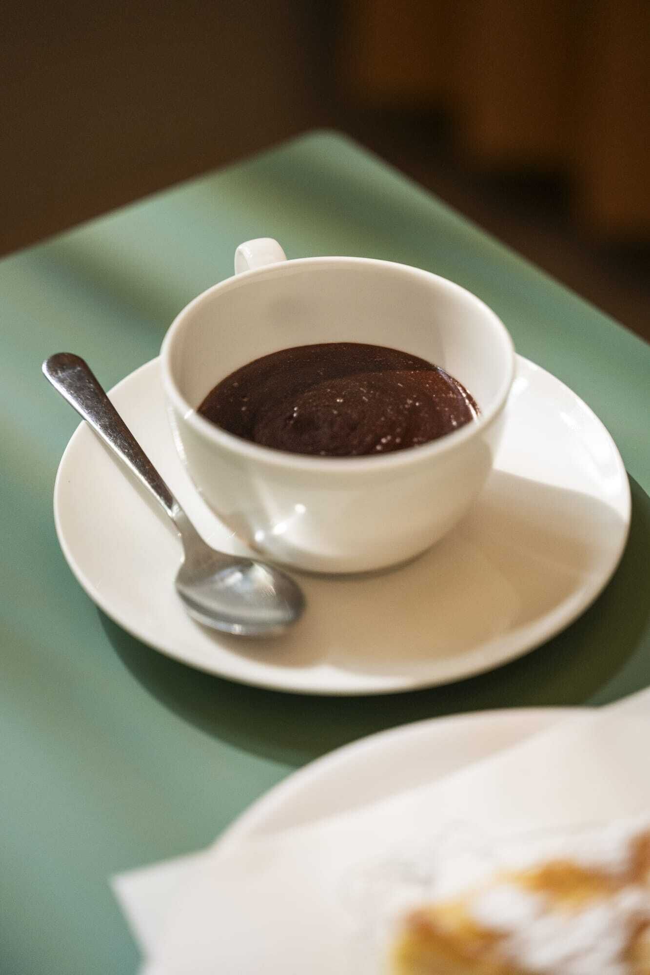 Una taza blanca con sopa oscura sobre un platillo blanco, con una cuchara, colocada sobre una mesa verde.