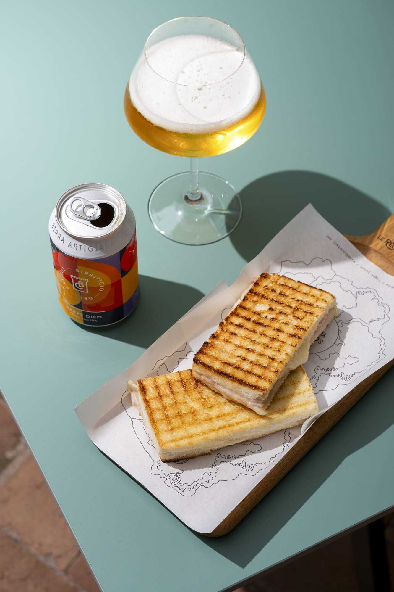 Una lata de cerveza, un vaso de cerveza y un sándwich a la parrilla sobre una mesa.