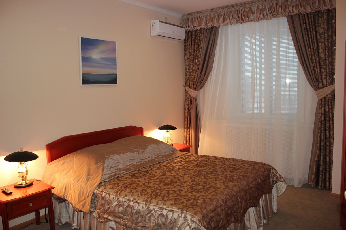 A neatly arranged bedroom with a bed, two bedside tables with lamps, an air conditioning unit, and a window with curtains.