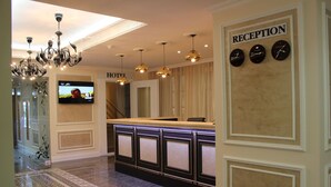 A hotel reception area with a reception desk, a chandelier, a television displaying a program, and a wall with clocks and a sign that reads "RECEPTION".