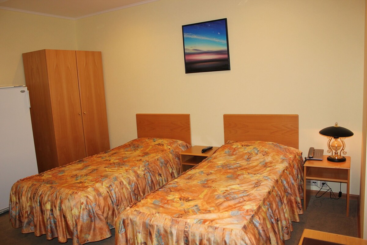 A room with two single beds, a wooden wardrobe, a desk with a lamp, and a painting on the wall.