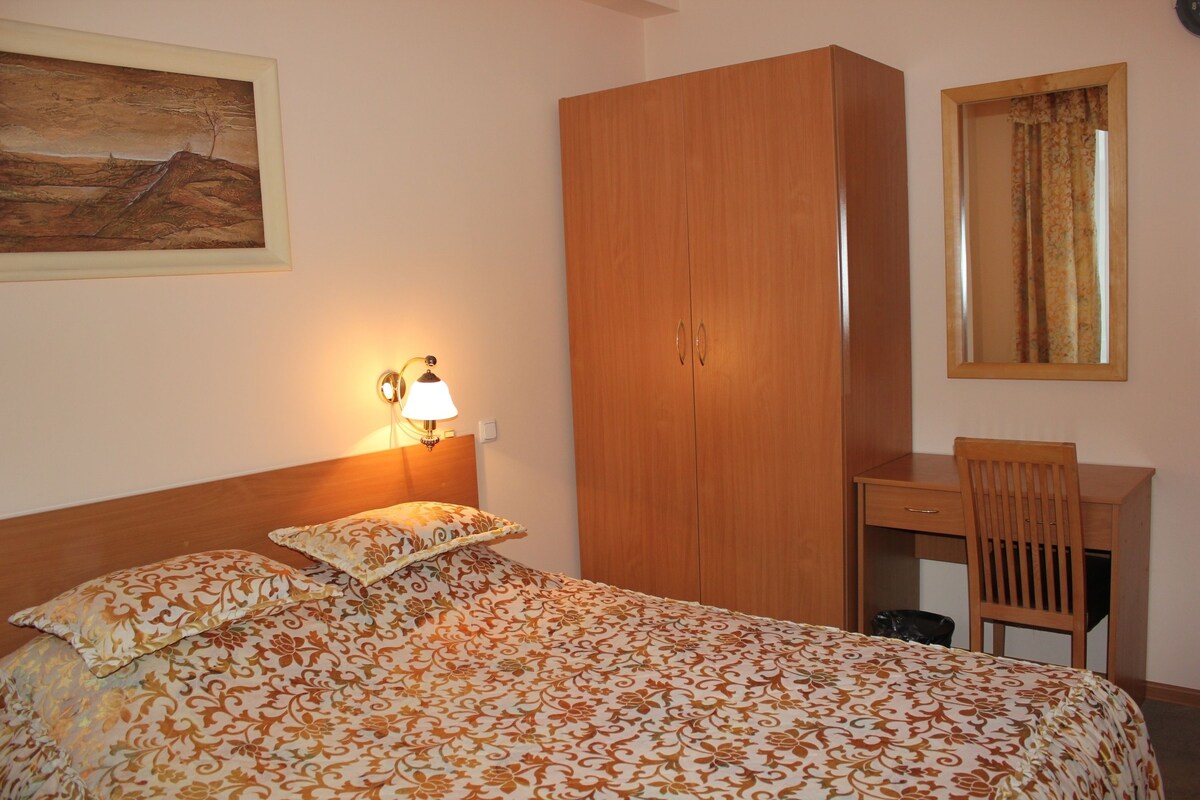 A bedroom with a bed, a wooden wardrobe, a desk with a chair, and a painting on the wall.