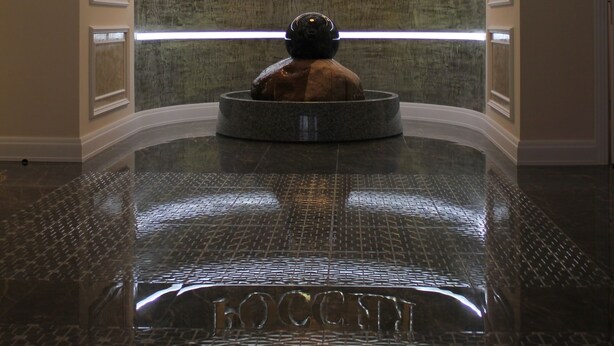 A lobby with a central circular seating area, a textured wall with illuminated text, and a reflective floor.