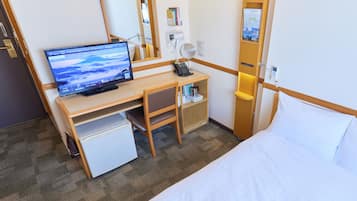 Ein Hotelzimmer mit einem Bett, einem Schreibtisch mit Computer, einem kleinen Kühlschrank, einem Nachttisch mit Lampe und einem Spiegel.
