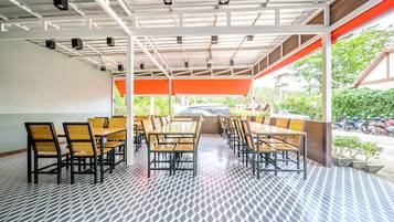 Un espacio cubierto para comer al aire libre con varias mesas y sillas, piso estampado y vistas a motocicletas estacionadas y vegetación.