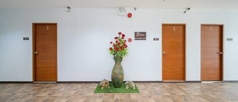 Un couloir avec trois portes en bois, chacune portant un numéro. Un tapis vert, sur lequel sont posés un vase rempli de fleurs rouges et deux petites sculptures, est placé au centre.