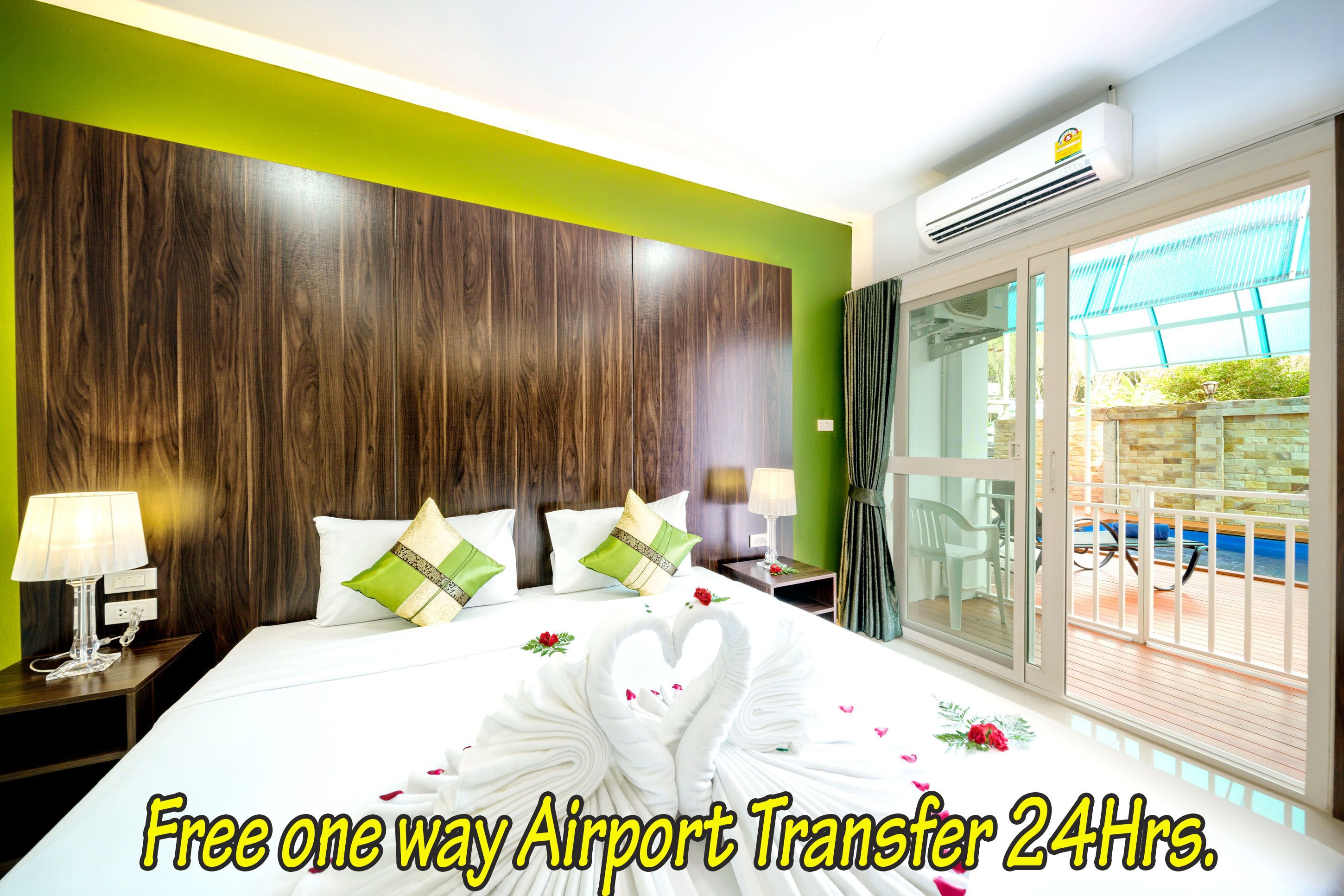 Una habitación de hotel con cabecera de madera, cama con sábanas blancas, almohadas verdes y una disposición de toallas con forma de corazón. Se ve una ventana con vistas a una piscina y un balcón. Se puede leer el texto "Free one way Airport Transfer 24Hrs.".