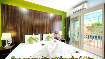 Una habitación de hotel con cabecera de madera, cama con sábanas blancas, almohadas verdes y una disposición de toallas con forma de corazón. Se ve una ventana con vistas a una piscina y un balcón. Se puede leer el texto "Free one way Airport Transfer 24Hrs.".