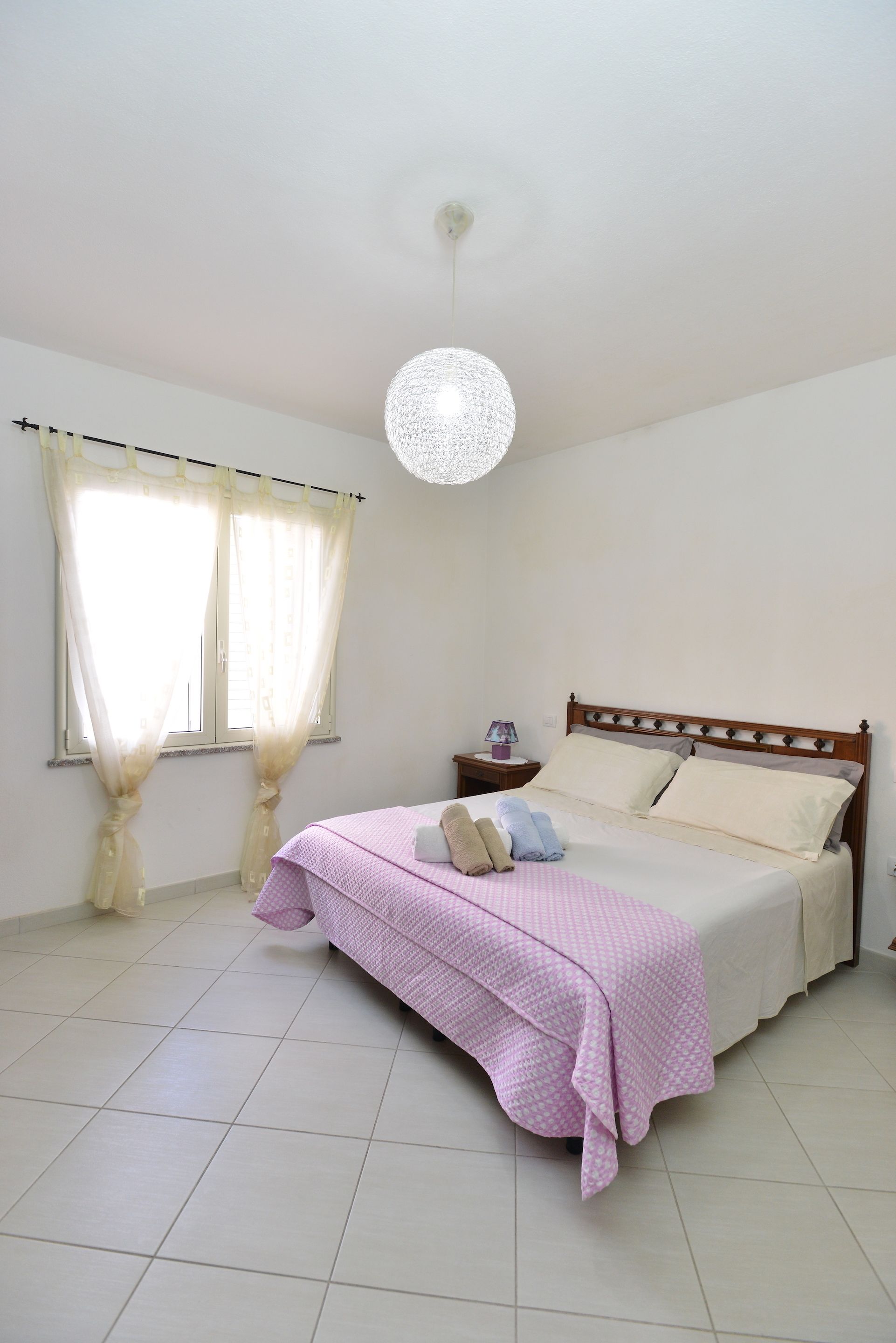 Een slaapkamer met een bed, gordijnen, een nachtkastje en een hanglamp.